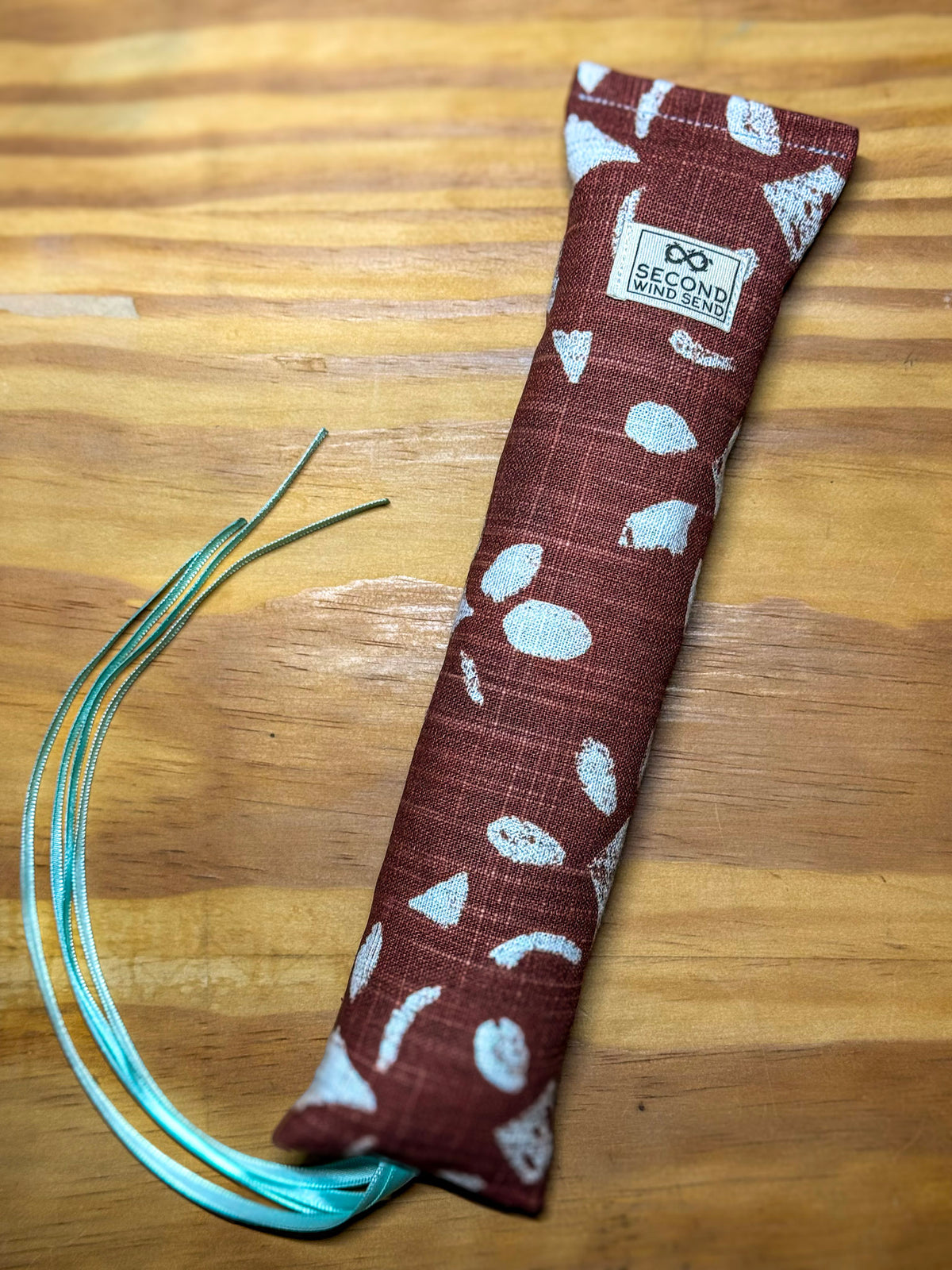 A fabric pouch or sleeve with a brown and white pattern, placed diagonally on a wooden surface. A small sewn label reads 'SECOND HAND STORES,' and several thin teal cords extend from one end.