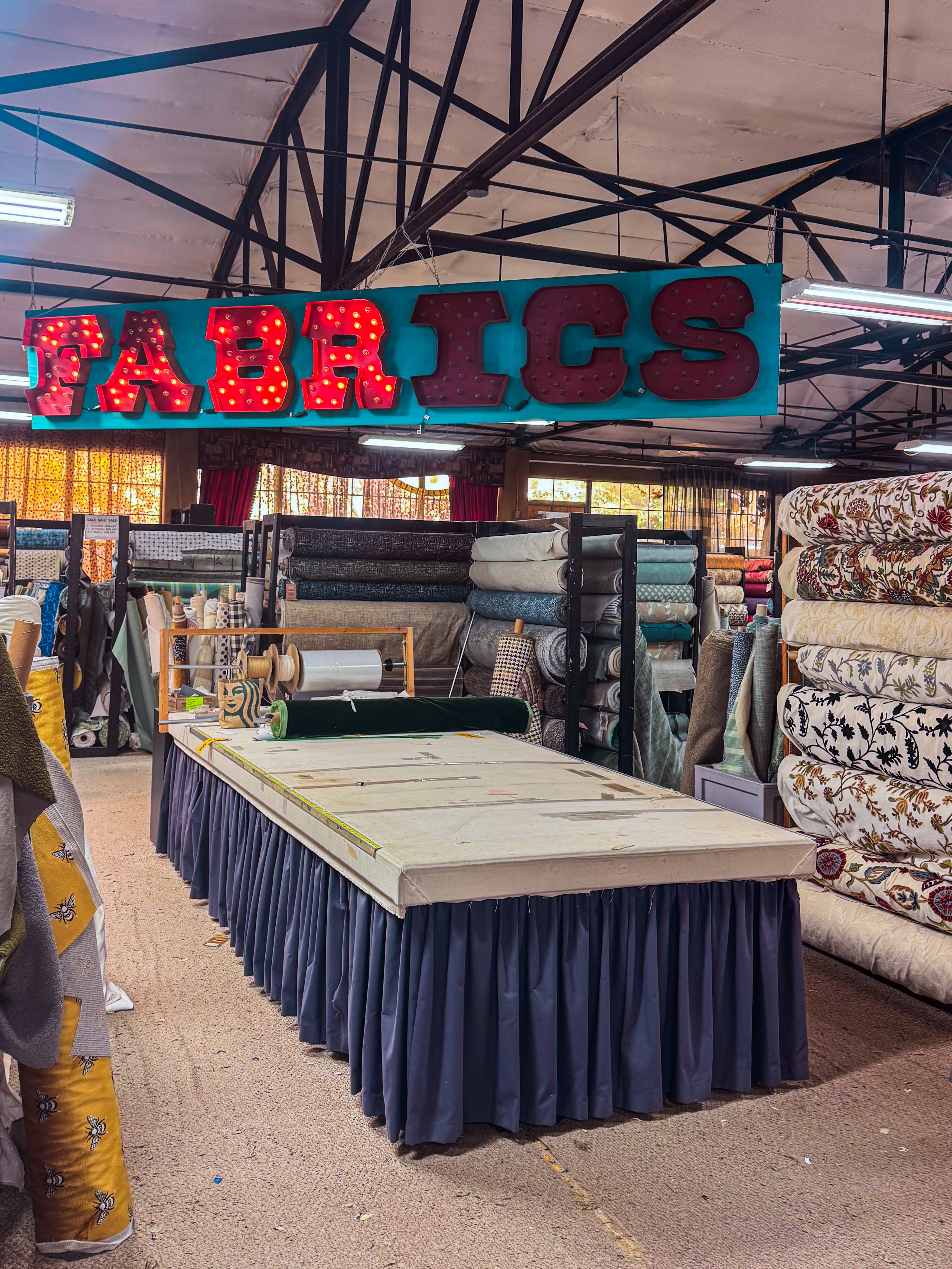 A large fabric store interior with a prominent red LED sign reading 'FABRICS' hanging above a central table draped in navy blue fabric. Stacks of folded and rolled textiles are visible on both sides, with the store's industrial ceiling and metal beams in the background.