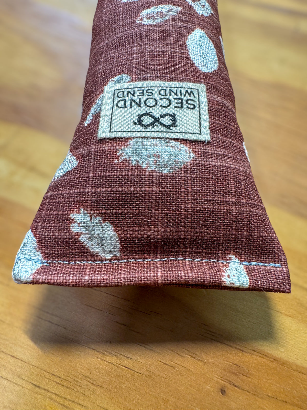 A close-up of a Second Wind Scent diffuser bag made of textured burgundy fabric with a white leaf pattern, resting on a wooden surface. The brand label is visible on the front.