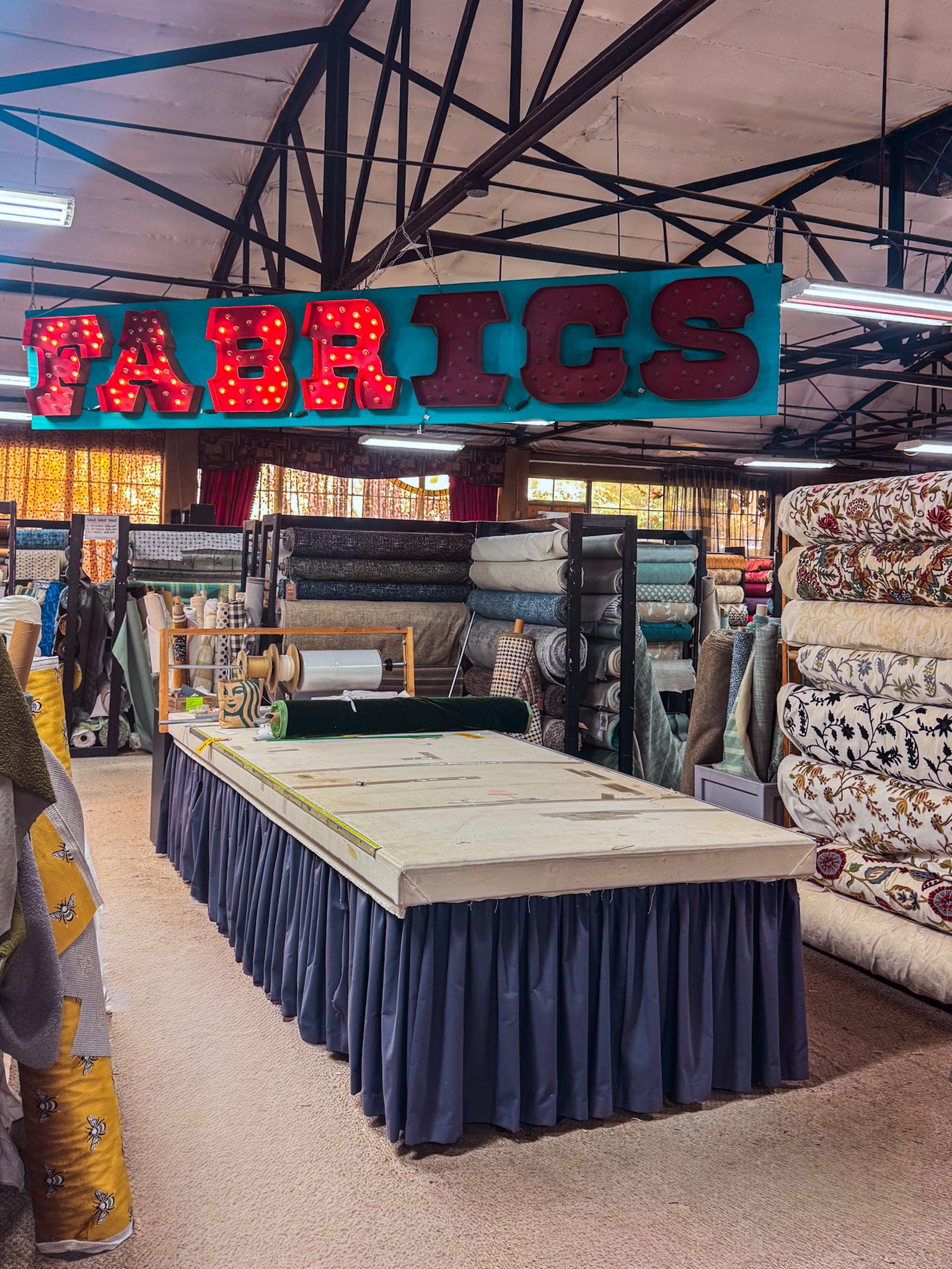 A large red LED marquee sign reading 'FABRICS' hangs above a display table in a fabric showroom. Rolled textiles, patterned fabrics, and sewing materials are visible on racks and shelves in the background under a high industrial ceiling.